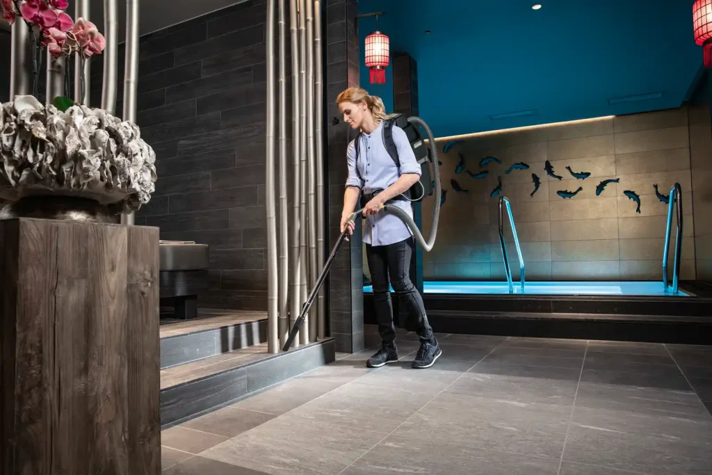 A woman wearing a backpack vacuum cleans tiled floors and steps in a modern spa or wellness area, with a pool, decorative lighting, and contemporary interior design in the background.