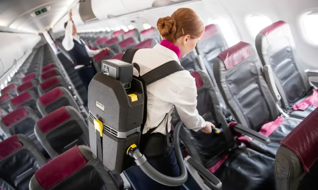 A person wearing a backpack vacuum cleaner cleans seats inside an airplane cabin, vacuuming between rows of empty passenger seats.