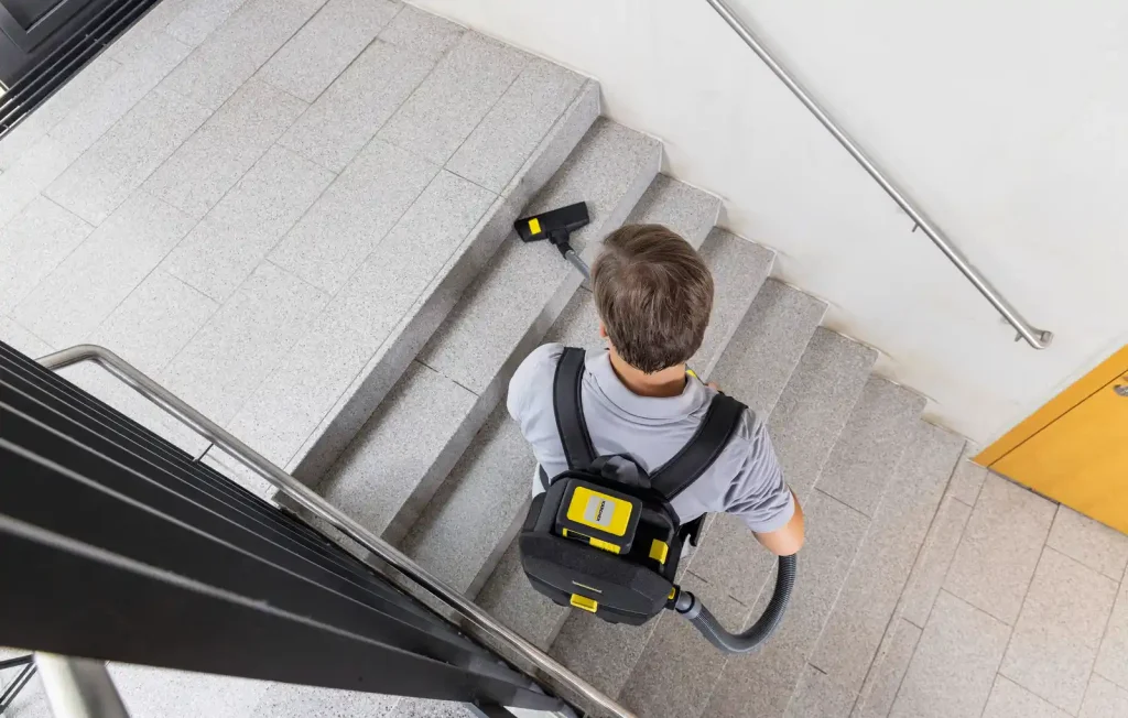 Overhead view of a person wearing a backpack vacuum cleaner while cleaning a tiled indoor staircase with a vacuum hose.