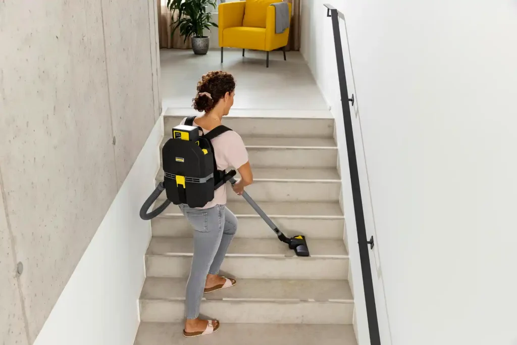 A person using a backpack vacuum cleaner to clean concrete stairs inside a modern building, vacuuming each step while standing on the staircase.
